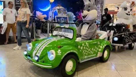 Mini Fusca verde exposto em um shopping, com personagens fantasiados ao fundo