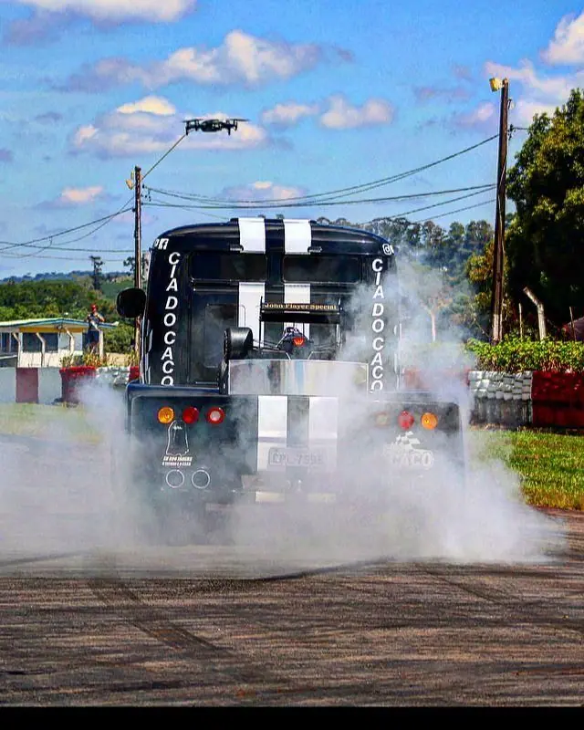 Caminhão preto e branco em manobra, envolto em fumaça, com um drone ao fundo
