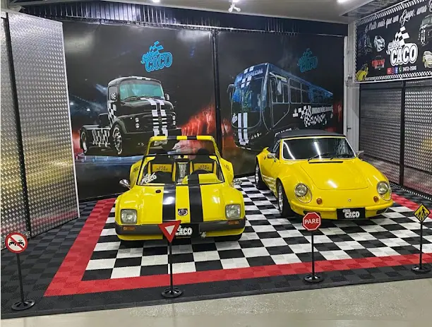 Dois carros esportivos amarelos sobre piso quadriculado, com painéis de caminhão e ônibus ao fundo