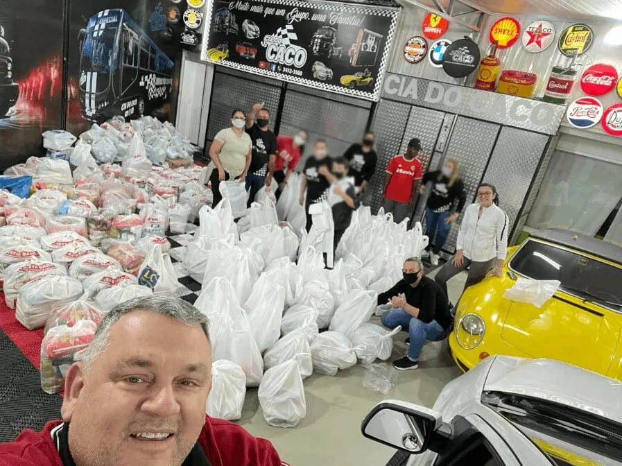 Voluntários em torno de sacolas de doação no galpão, com um homem fazendo selfie em primeiro plano