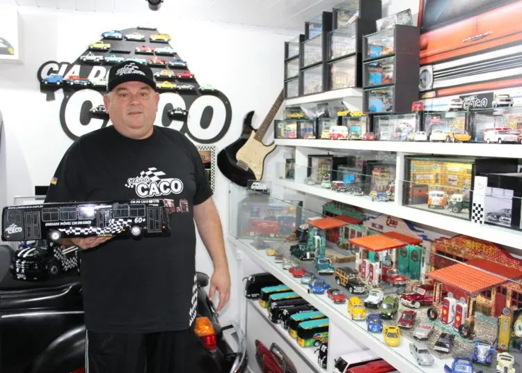 Homem de boné e camiseta da Cia do Caco segura uma miniatura de caminhão diante de estantes de miniaturas
