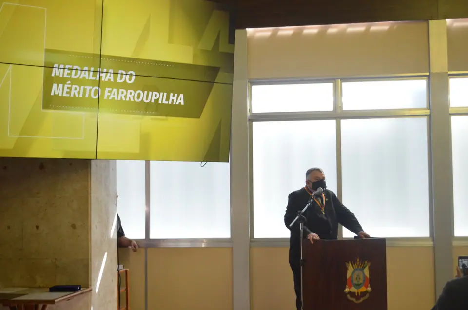 Homem discursa em uma tribuna diante de um painel com o texto Medalha do Mérito Farroupilha
