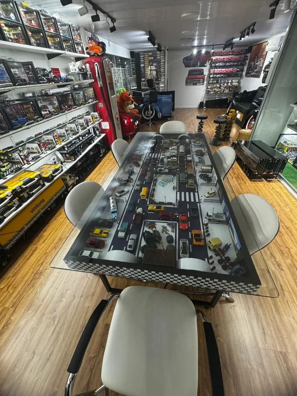 Mesa de vidro com uma maquete de cidade e miniaturas de carros, cercada por cadeiras brancas